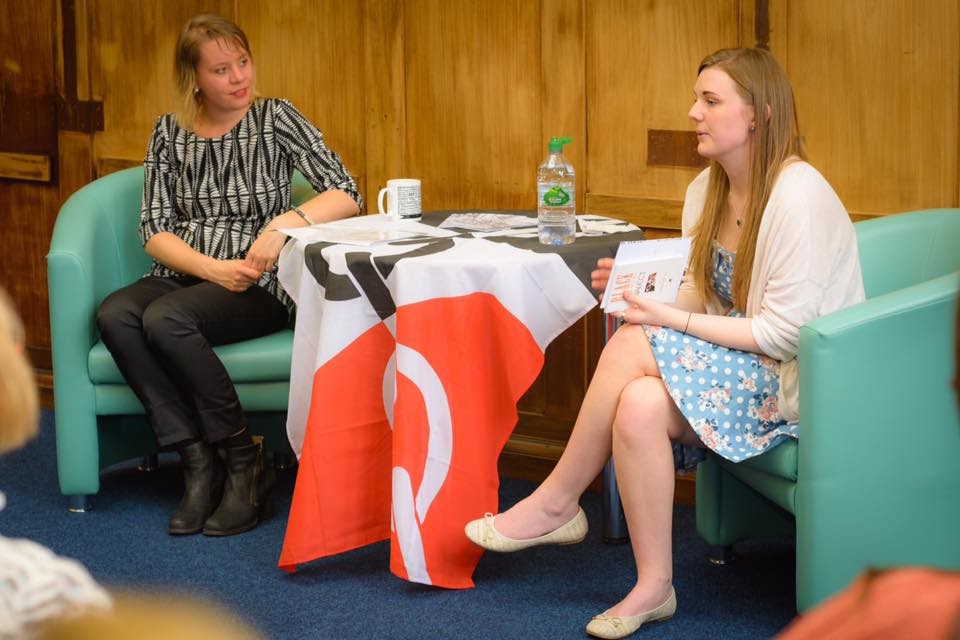 Two young women (Jorine and Nellie) sit on either side of a table, which is draped with a Black Country flag. Jorine is looking at Nellie, who is addressing a group of people out of shot.