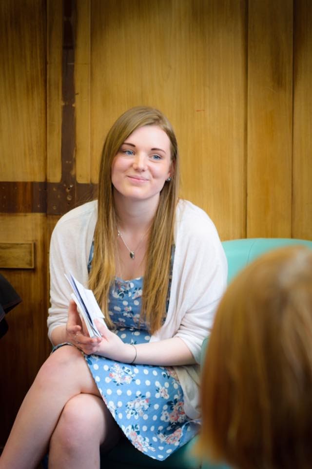 A young woman (Nellie), sits on a chair, smiling at a group of people out of shot.