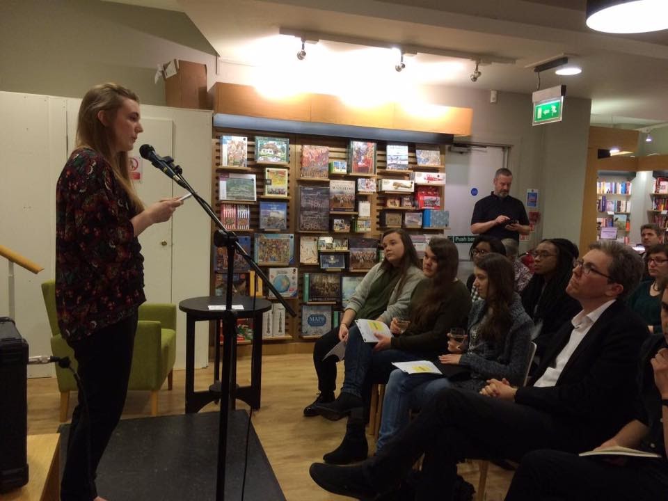 A young woman stands on a stage, behind a mic, reading to a room of seated people, only the first row can be seen. The backdrop is a bookshop.