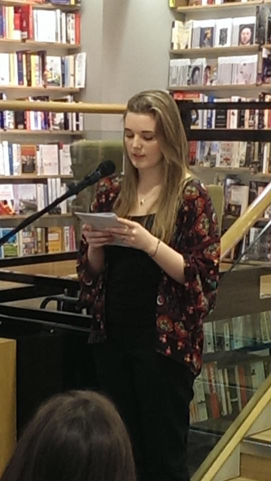 A young woman reads from behind a mic, holding a booklet in her hands. The backdrop is a bookshop.