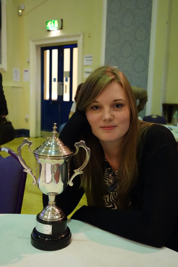A young woman is sat, semi-smiling at the camera, her jaw resting on her hand. On the table in front of her there is a large silver trophy.