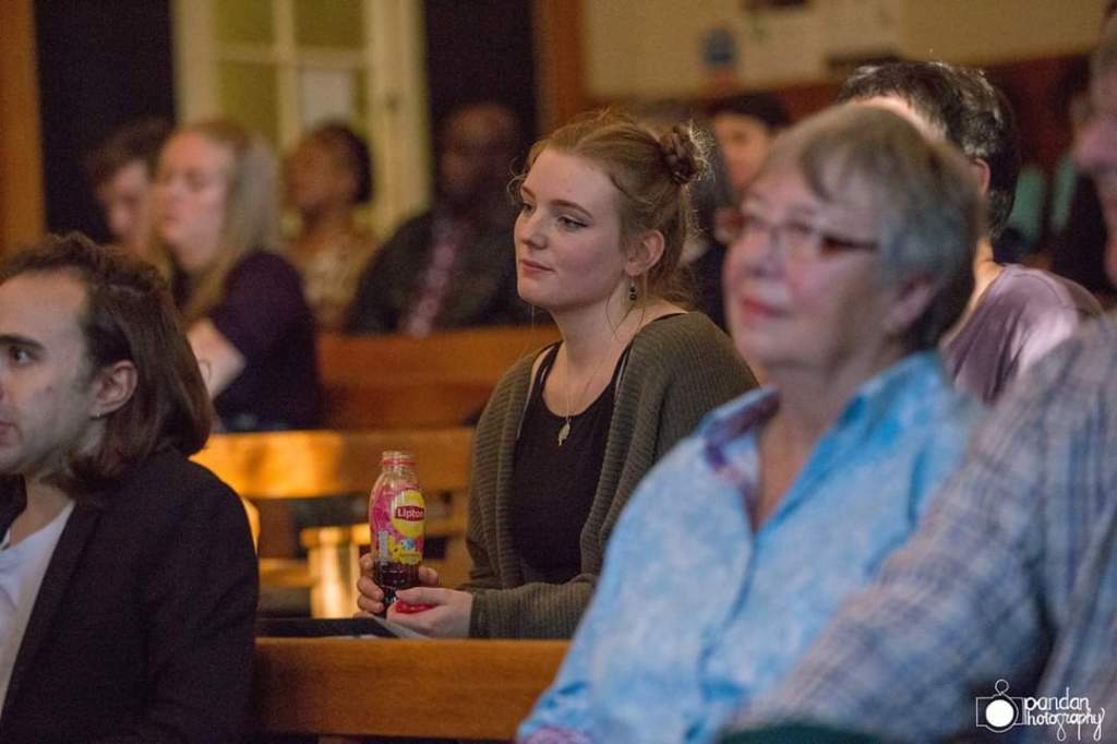 A lecture theatre filled with people looking up (at a reader on stage, not pictured). The focus is on one young woman (me) listening and smiling.