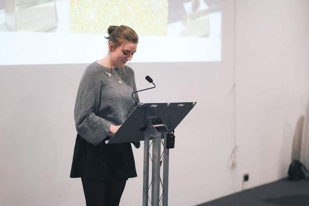 A young woman stands behind a lecturn, looking down at what she is reading with a smile on her face.