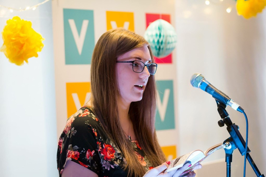 A young woman reads behind a mic. Behind her is a banner patterned with blocks of turquoise, yellow and pink, with a white V in the centre of each one.