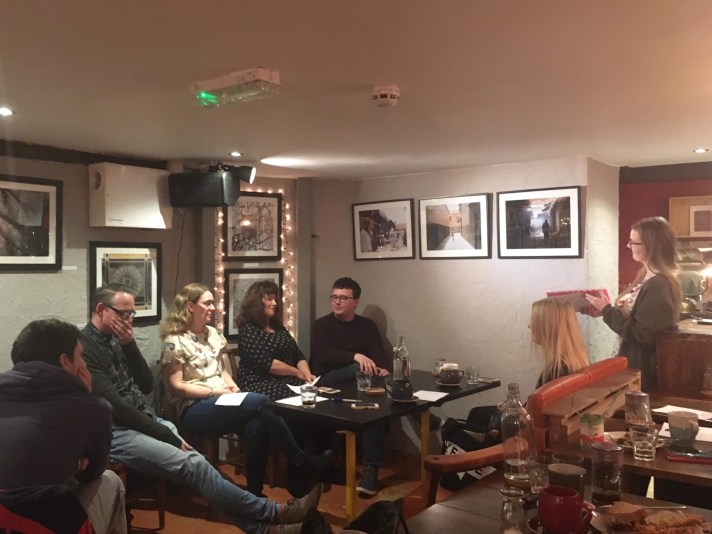 Four poets (two male, two female), sit in a row, behind a table. A young woman, the host, is standing facing them. Seated around are various spectators.
