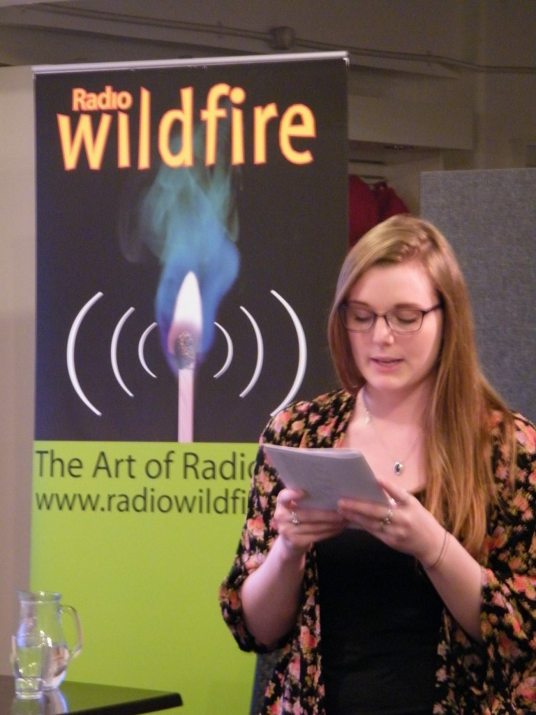 A young woman (Nellie) stands reading from a small booklet she holds in her hands. Behind her is a banner for 'Radio Wildfire'.