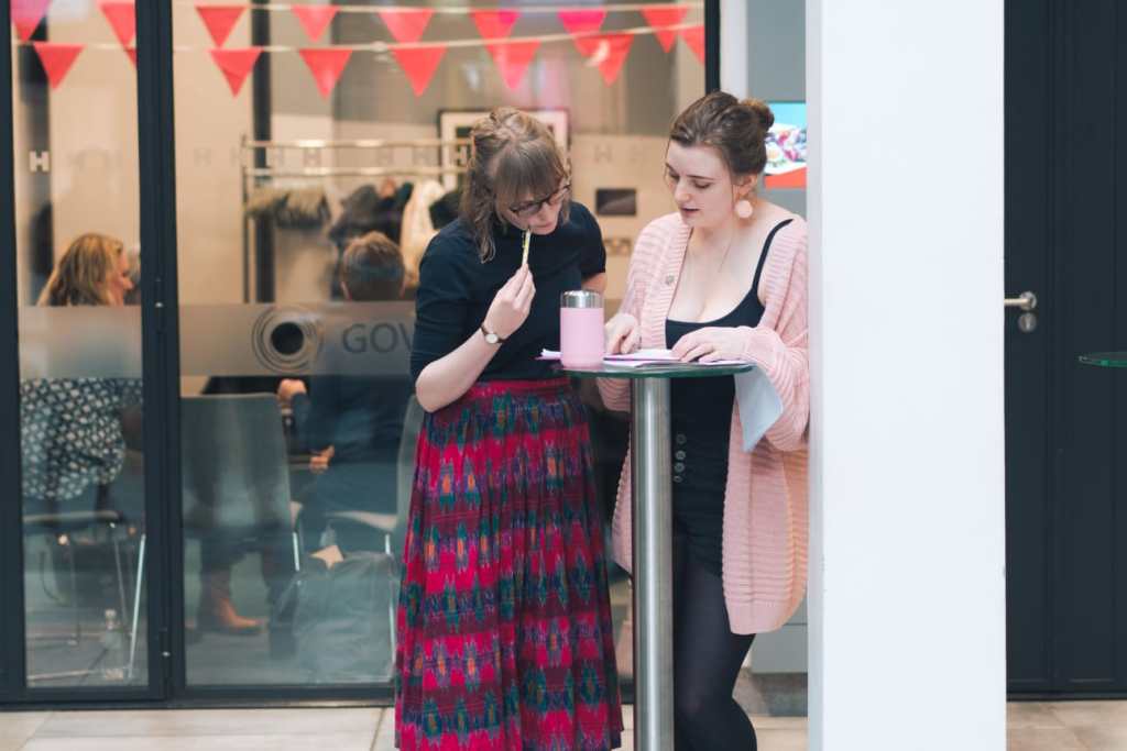 Two young woman stand behind a high table, looking down at some papers.