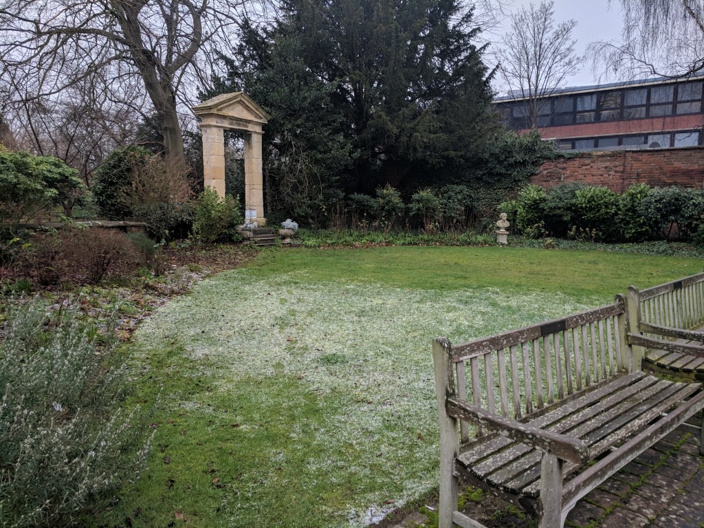 A triangular lawn, lightly dusted with frost. It is bordered by plants and trees, and a large stone archways at the apex.