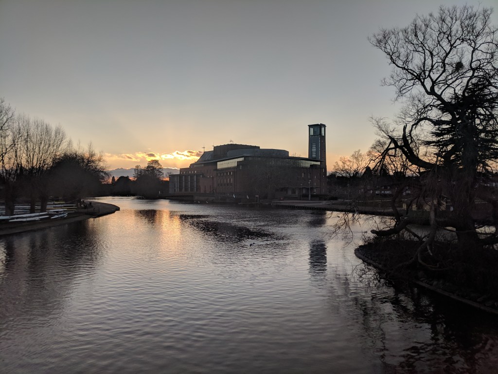 The Royal Shakespeare Theatre on the bank of the River Avon, silhouetted against a sunset sky.